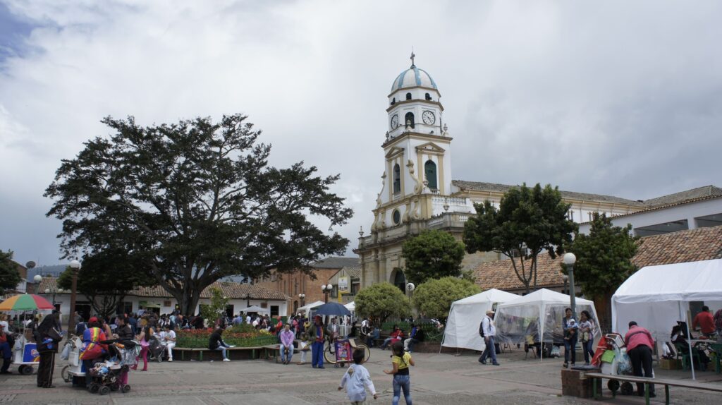Vista del centro de Chía, Colombia, con una iglesia colonial en el fondo, personas caminando y compartiendo en la plaza, rodeada de árboles y arquitectura tradicional.