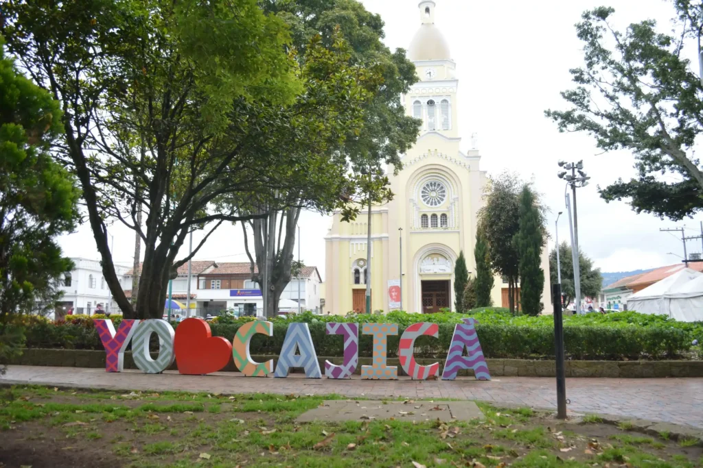 Imagen del parque principal de Cajicá, Cundinamarca, con la iglesia San Roque al fondo y el colorido letrero "Yo ❤️ Cajicá" en primer plano.